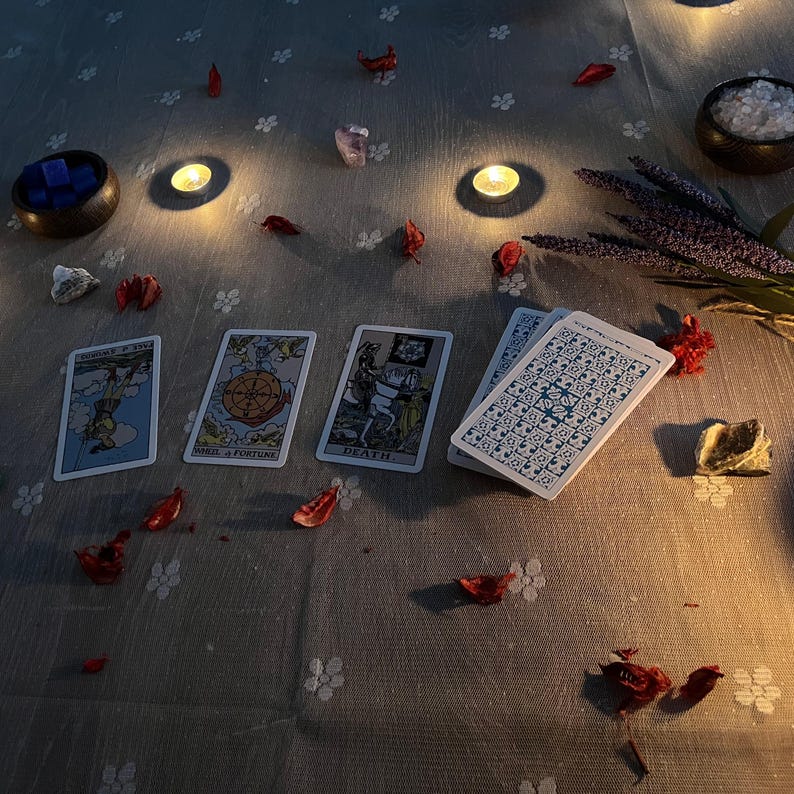 May include: A mystical arrangement featuring tarot cards with illustrations and text, scattered rose petals, candles, crystals, and a bowl of blue cubes. The cards are arranged on a grey floral patterned surface, creating a spiritual ambiance.