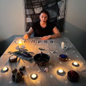May include: A woman is seated at a table with tarot cards, candles, and various items. The table is covered with a light grey cloth with a floral pattern. Items include bowls of salt, herbs, and crystals, and bundles of cinnamon sticks.