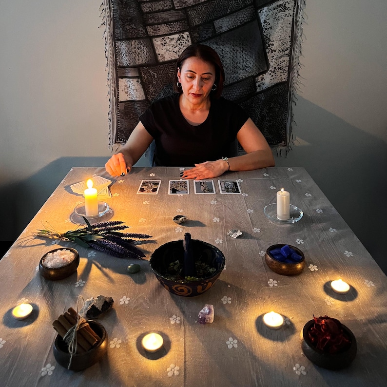 May include: A woman is seated at a table with tarot cards, candles, and various items. The table is covered with a light grey cloth with a floral pattern. Items include bowls of salt, herbs, and crystals, and bundles of cinnamon sticks.
