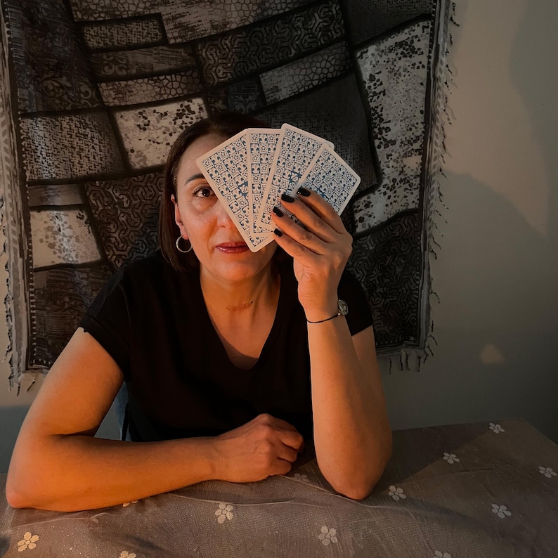 May include: A person holding a fan of tarot cards in front of their face. The cards have a white background with a blue and white pattern. The person is wearing a black top and has dark nail polish.
