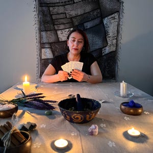 May include: A woman seated at a table, holding tarot cards. The table is set with candles, bowls of herbs and crystals, and other mystical items. The scene is dimly lit, creating a spiritual atmosphere.