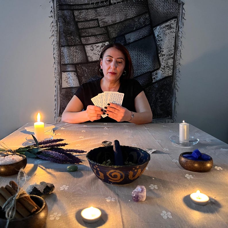 May include: A woman seated at a table, holding tarot cards. The table is set with candles, bowls of herbs and crystals, and other mystical items. The scene is dimly lit, creating a spiritual atmosphere.