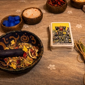 May include: A still life arrangement featuring a mortar and pestle with dried herbs, small wooden bowls with blue cubes and salt, a deck of tarot cards labelled "King of Pentacles", and a bundle of dried flowers.