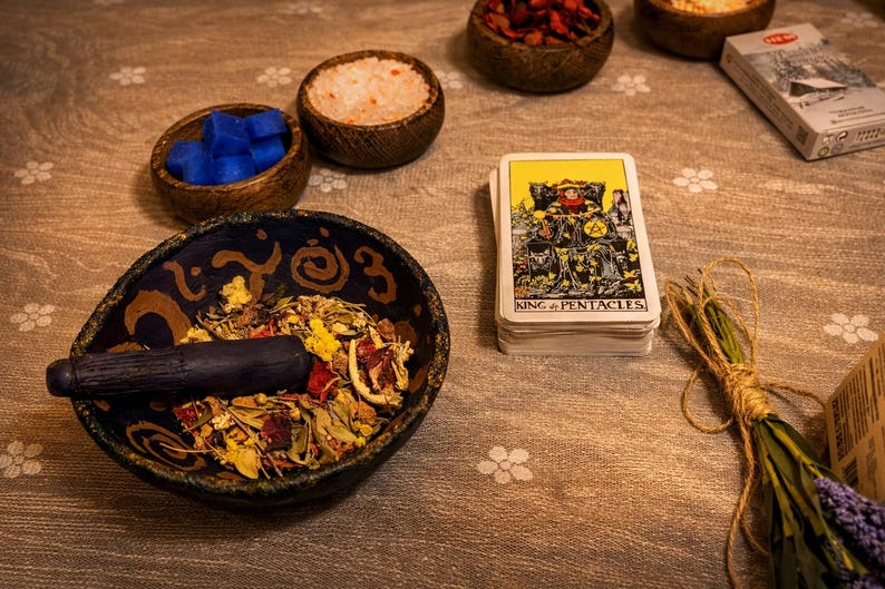 May include: A still life arrangement featuring a mortar and pestle with dried herbs, small wooden bowls with blue cubes and salt, a deck of tarot cards labelled "King of Pentacles", and a bundle of dried flowers.