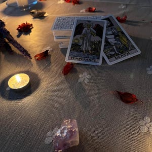 May include: A low-lit image showing tarot cards, a burning candle, and red petals. Two cards are visible, marked "Temperance" and "Death." An amethyst crystal and a deck of cards are also present, creating a mystical atmosphere.