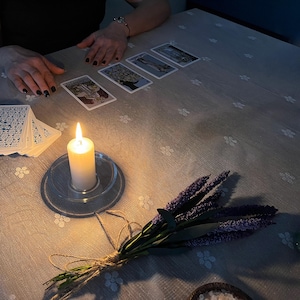 May include: A lit white candle in a clear glass holder sits on a floral patterned tablecloth, next to a deck of cards and several tarot cards. A bundle of lavender tied with twine rests nearby. The scene is dimly lit.