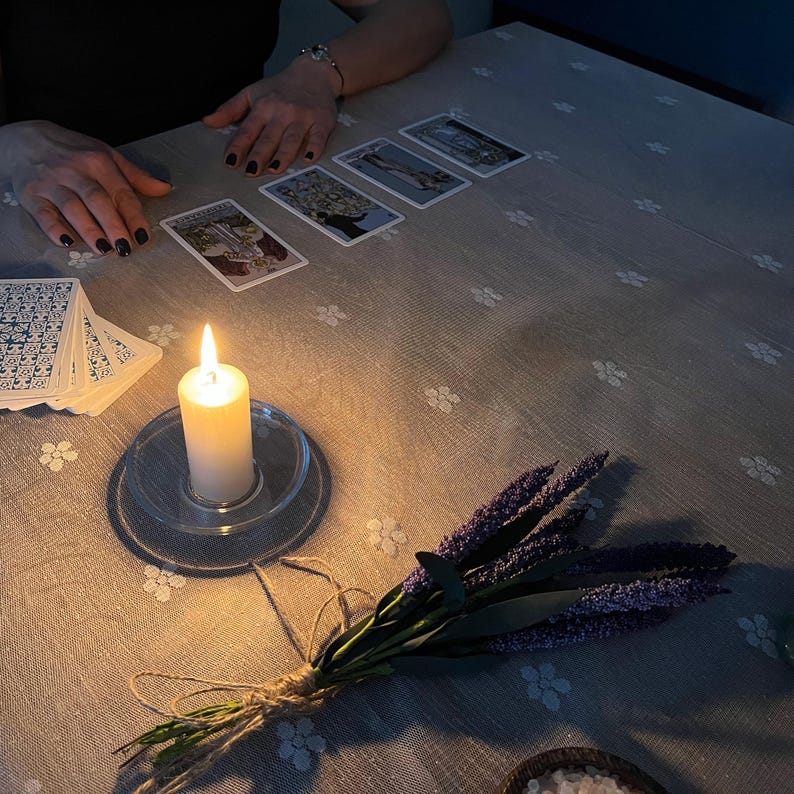 May include: A lit white candle in a clear glass holder sits on a floral patterned tablecloth, next to a deck of cards and several tarot cards. A bundle of lavender tied with twine rests nearby. The scene is dimly lit.