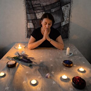 K&ouml;nnte beinhalten: Eine Person mit gefalteten H&auml;nden betet an einem Tisch mit Kerzen, Kristallen und kleinen Schalen. Der Tisch ist mit einem hellgrauen Tuch bedeckt. Im Hintergrund h&auml;ngt ein Wandteppich. Die Szene wird durch Kerzenlicht beleuchtet und erzeugt eine mystische Atmosph&auml;re.