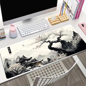 May include: A long rectangular desk mat featuring a black and white Asian-inspired landscape design. The artwork depicts a traditional building, mountains, and a flowering tree. The mat is placed on a white desk with a computer and keyboard.
