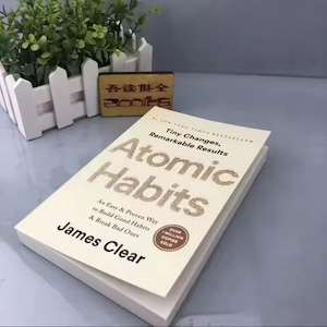 May include: A hardcover book titled "Atomic Habits" by James Clear. The book has a cream-colored cover with the title in large, glittery letters. The book is open, revealing the title and subtitle, with a sticker indicating over a million copies sold.