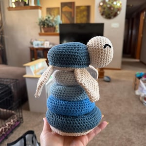 May include: Crocheted turtle stacking toy in shades of blue and beige. The turtle has a stack of four graduated rings, with a beige head and flippers. The toy is handmade and appears soft and cuddly.