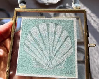 May include: A square brass frame with a light blue velvet ribbon hanger containing a square piece of textured paper. The paper features a white illustration of a scallop shell on a sage green background with the initials MG in the bottom corner.