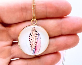 May include: A gold-toned circular pendant necklace featuring a detailed illustration of a spotted seashell with pink and brown hues on a white background. The pendant hangs from a delicate gold-toned chain, showcasing a clear, smooth finish.