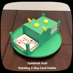 May include: A green SpinDeck Dual rotating 2-bay card holder with yellow details. The card holder has two sections for holding playing cards, with a deck of cards visible in one bay. The text "SpinDeck Dual Rotating 2-Bay Card Holder" is at the bottom.