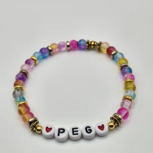 May include: A beaded bracelet featuring multicolored, translucent, crackle-textured beads interspersed with small gold-toned metal spacers. The center of the bracelet displays white circular beads spelling the name PEG, flanked by two red heart icons.