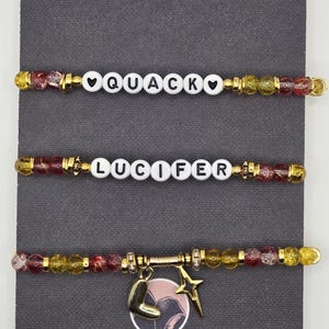 May include: Three beaded bracelets with white letter beads spelling "QUACK" and "LUCIFER". The bracelets have gold accents and charms, including a star and a letter "L". The beads are a mix of red, yellow, and clear.