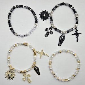 Op de afbeelding: Vier armbanden met kralen en bedels. Twee armbanden zijn zwart en grijs met kruisen, een doodskist en een zonne-uitbarsting bedel. De andere twee zijn wit en goud met een kruis, doodskist en zonne-uitbarsting bedel. Eén armband spelt "MON CHER".