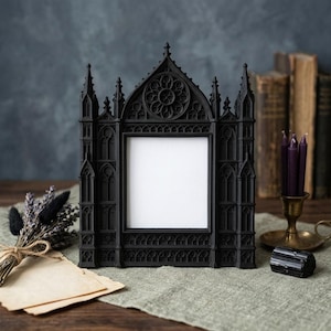 May include: Black gothic-style picture frame with intricate architectural details, including spires and a rose window design. The frame has a blank white space for a photo. The frame is on a wooden surface with other gothic-themed decor.