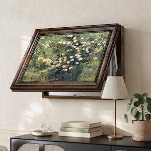 Puede incluir: Un armario de pared de madera oscura con un panel frontal abatible que muestra un estampado de rosas blancas en un jardín. Debajo del armario hay una lámpara de mesa con pantalla blanca, tres libros apilados y un pequeño plato blanco.