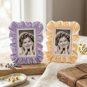 May include: Two decorative photo frames, one lavender and one pale yellow, each with a ruffled satin-like border. The frames hold black and white portraits of a smiling child. The frames are displayed on a wooden surface.
