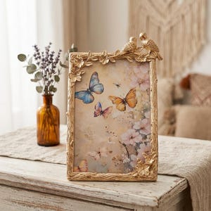 Può includere: Una cornice dorata con un design a farfalle e fiori. La cornice ha una forma rettangolare ed è adornata con fiori scolpiti e due piccole figure di uccelli nella parte superiore. L'immagine all'interno presenta farfalle e fiori.