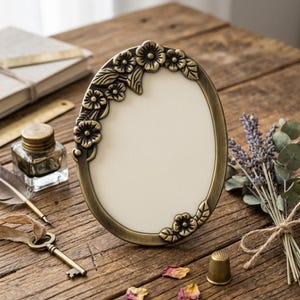 Peut inclure: Un cadre photo ovale de style antique avec un motif floral. Le cadre est en métal de couleur bronze et possède un insert de couleur crème. Le cadre est entouré d'autres objets vintage, dont une clé, un encrier et des fleurs séchées.
