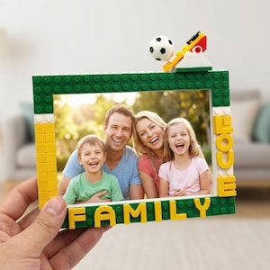 Könnte beinhalten: Ein rechteckiger Bilderrahmen aus grünen, gelben und weißen Kunststoff-Bausteinen. Der Rahmen zeigt unten das Wort FAMILY in gelben Buchstaben, rechts das Wort LOVE und oben ein kleines Spielzeug-Fußballmodell als Dekoration.