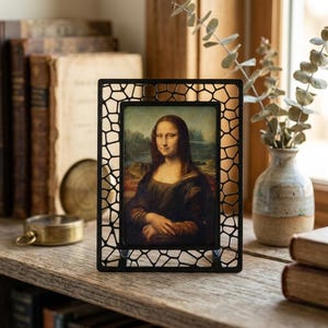 May include: A black metal picture frame with a mosaic-like design surrounds a print of the Mona Lisa painting. The frame is displayed on a wooden shelf with antique books and a small vase.
