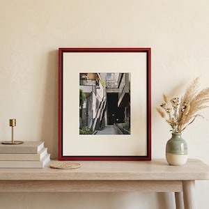 May include: A framed photograph with a red wooden frame, showcasing an architectural scene. The photograph is displayed on a light-colored wooden table, alongside a stack of books, a candle holder, a coaster, and a vase with dried flowers.
