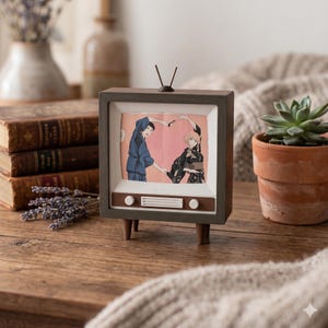 May include: A miniature wooden television with a screen displaying a romantic anime scene. The TV has a dark brown frame, a light brown speaker panel, and two white knobs. The scene shows two anime characters forming a heart shape with their arms.