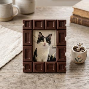 Puede incluir: Un marco de fotos rectangular con un acabado marrón oscuro moldeado para parecerse a una tableta de chocolate segmentada. El marco contiene una fotografía de un gato blanco y negro con ojos verdes, frente a una pared clara.