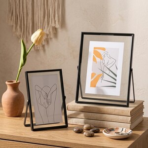 Puede incluir: Dos marcos de fotos de metal negro con vidrio transparente, cada uno con impresiones artísticas. Un marco descansa sobre una pila de libros. Un jarrón con un tulipán amarillo y un pequeño plato decorativo también están sobre la superficie de madera.