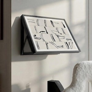 May include: A rectangular wall-mounted display shelf with a black frame, featuring a tilted surface that holds a grid of square tiles. Each tile displays a cream background with abstract black brushstroke patterns arranged in a repeating sequence.
