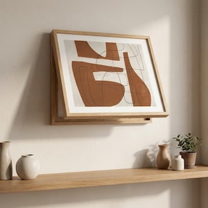 Könnte beinhalten: Abstrakter Kunstdruck in einem hellen Holzrahmen. Das Kunstwerk zeigt terrakottafarbene Formen und schwarze Strichzeichnungen auf cremefarbenem Hintergrund. Das gerahmte Kunstwerk ist an einer weißen Wand montiert, darunter ein Holzregal.