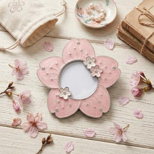 May include: A pink flower-shaped picture frame with a silver border and small decorative flowers. The frame is adorned with rhinestones and sits on a white wooden surface, surrounded by cherry blossoms and petals. A small bag and a dish with pearls are also present.