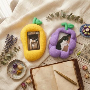 May include: A yellow mango-shaped photo frame and a purple grape-shaped frame, each holding a black and white cat photo. The frames are on a cream-colored surface, with a journal, pen, and decorative items.