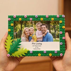 Könnte beinhalten: Ein rechteckiger Bilderrahmen aus grünen Bausteinen, verziert mit kleinen gelben und rosa Blumen sowie zwei großen hellgrünen Blattelementen. In der Mitte ist ein Familienfoto mit dem Text Pin Living - DIY Photo Frame - 10 cm x 15 cm zu sehen.