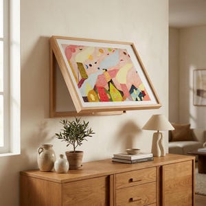 Könnte beinhalten: Ein holzgerahmtes Kunstwerk mit einem abstrakten Design in Rosa, Gelb, Blau und Grün hängt an einer Wand. Der Rahmen ist teilweise geöffnet. Unter dem Kunstwerk befindet sich ein Holzschrank mit einer Lampe, Büchern und dekorativen Gegenständen.