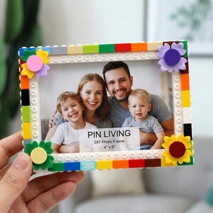 Könnte beinhalten: Ein rechteckiger Bilderrahmen aus bunten Kunststoff-Bausteinen mit einem weißen Innenrand und dekorativen blumenförmigen Akzenten an den Ecken. Der Rahmen zeigt ein 4" x 6" Foto einer Familie mit dem aufgedruckten Text PIN LIVING.