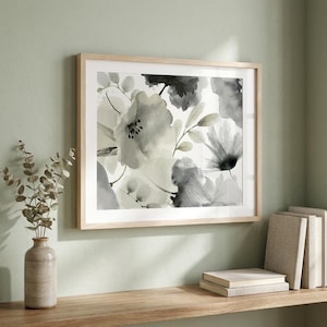 May include: A framed floral art print featuring a watercolor design of large, abstract flowers in shades of gray and white. The artwork is displayed in a light wood frame, creating a neutral and elegant aesthetic. The print is displayed on a wooden shelf.