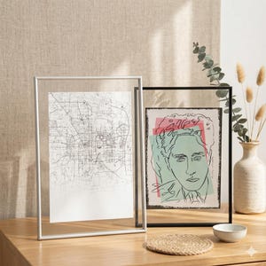 May include: Two framed art prints. One shows a detailed black and white map, the other a colourful illustration of a person's face with pink, green, and black accents. Both are displayed on a wooden surface.