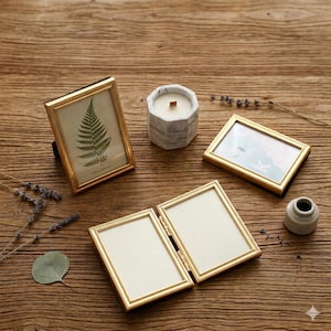 Puede incluir: Una colección de elementos decorativos sobre una superficie de madera. Incluye marcos dorados, un portavelas con diseño de mármol con una vela encendida y un pequeño jarrón blanco. Un marco muestra una impresión de helecho.