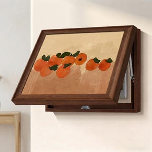 May include: A rectangular wall-mounted cabinet with a dark brown wooden frame. The front panel features a horizontal painting of several orange fruits with green leaves against a light tan background. The cabinet is hinged to open outwards.
