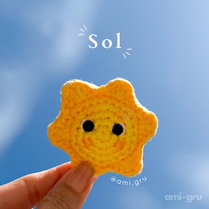 Puede incluir: Una aplicación de sol tejida a ganchillo en color amarillo brillante con un centro circular texturizado y cinco puntas redondeadas. El sol tiene dos ojos negros pequeños y redondos, con el texto Sol arriba y el nombre ami.gru debajo.