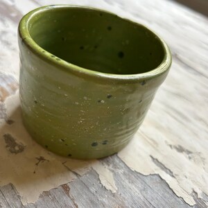 Könnte beinhalten: Eine handgefertigte, grüne Keramiktasse mit gesprenkelter Oberfläche. Die Tasse hat eine zylindrische Form mit einem leicht ausgestellten Rand. Die Außenseite ist meliert grün mit dunkleren grünen und schwarzen Sprenkeln. Die Tasse wird wahrscheinlich zum Trinken von Getränken verwendet.