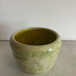 May include: A small, round ceramic cup with a glossy finish. The exterior features a mottled, light green and cream-colored glaze with subtle horizontal ridges. The interior of the cup is a smooth, vibrant yellow-green hue with a reflective surface.