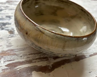 May include: A handmade ceramic bowl with a unique, organic shape. The bowl features a speckled, earthy color palette with shades of beige, brown, and cream. The rim has a darker brown glaze. This piece is ideal for serving or as a decorative accent.