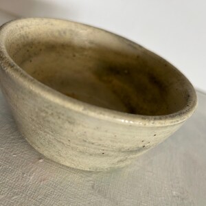 May include: A small, round ceramic bowl with a speckled, off-white and beige glaze. The bowl features a smooth, slightly tapered exterior and a wide, open interior with visible dark brown and grey flecks throughout the textured surface.