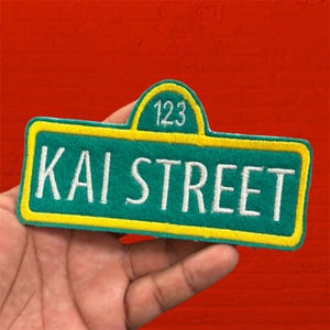 May include: A rectangular embroidered patch with a rounded top section, featuring a teal background and a yellow border. The text "KAI STREET" is stitched in white across the center, with the number "123" embroidered in white at the top.