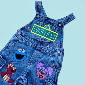 May include: Blue denim overalls featuring embroidered patches of Cookie Monster, Elmo holding a gift, and Abby Cadabby. A green street sign patch reads LUCILLE ST 123, surrounded by small pink and yellow embroidered flower outlines.